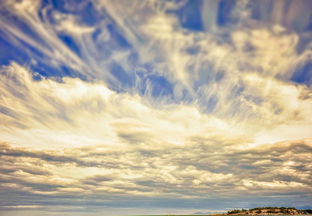 White streaky clouds hovering over a little island. The sky looks huge compared to the land mass. The dramatic clouds appear to hold the sun as they are illuminated from within causing them to have a yellowish hew. The sky is deep blue up top and grey blue below.
