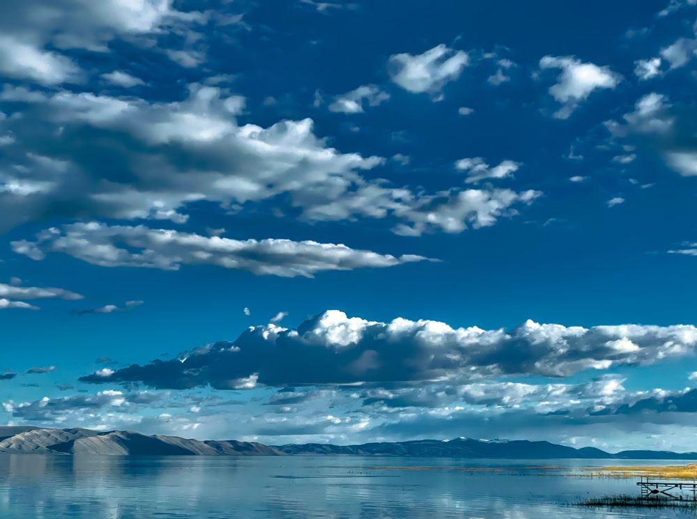 Looking outward at a lake to the opposite shore. A small yellow toned grassy patch is right of frame accompanied by a small wooden dock. There are long fluffy clouds floating in the deep blue sky and throwing shadows on the lake.