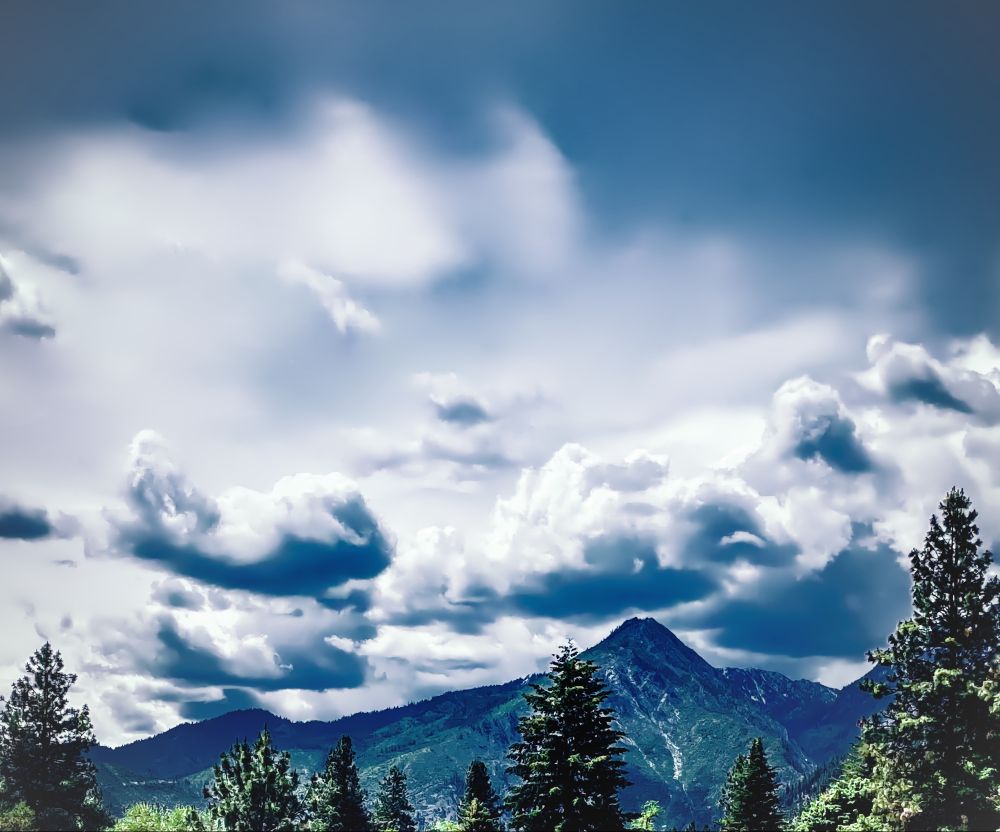 Eastern Washington mountain taken from Leavenworth on a spring day. Evergreen tree tops frame the bottom of the shot looking outward to a distant mountain. Dark clouds are smattered around the mountain top with a deep grey blue sky above.