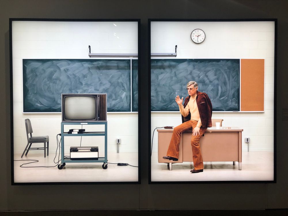 Rodney Graham’s Media Studies ‘77 at the Royal Ontario Museum in Toronto, depicting the artist sitting on a desk as if about to present a lecture. 