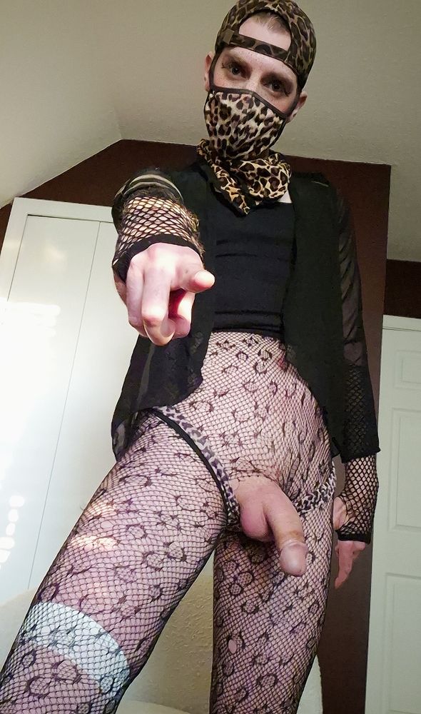 HylianTwink (a fair-skinned nonbinary person with bright blue eyes, red-brown hair, and freckles wearing a leopard-print baseball hat, mask, bandana, boyshorts, sheer black shirt over a black tank top, and fishnet gloves and tights) standing above and pointing down at the camera with their boyshorts pulled below their thick soft cock to let it hang between their thighs.