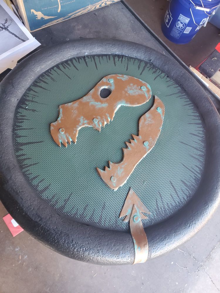 A round shield made of foam over a bare workshop floor. It has a black rounded edge, and it's face features a flat metallic cut-out of a wolf skull with open jaw in copper with bolts and edges distressed with green verdigris. The surface of the shield has a tatami texture and is painted forest green. The black color of the rim extends onto the green surface in uneven spikes 1-3 inches long, reminiscent of a silhouetted fir treeline.