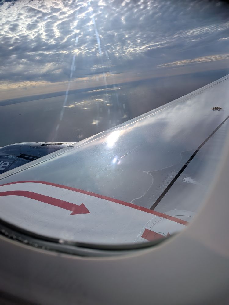 A picture of a plane wing from a different angle than the last takes up most of the view. Some white clouds scud overhead but you can see a bit of the ocean beyond the wing.