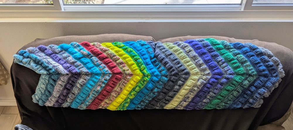 Small Street Shawl by Noma Ndlovu (BiggerThanLife) on Ravelry. Cashmere / merino blend from 2025 Coraline advent from YarnBaby. A multicolored scarf with 24  Vs pointing in one direction. The fabric has an attractive puckered appearance due to changing needle sizes and sharp increases and decreases in each stripe. There are prominent bright red and yellow stripes, but the scarf is dominated by blues and purples, both variegated and tonals with colors smoothly transitioning from one stripe to the next.