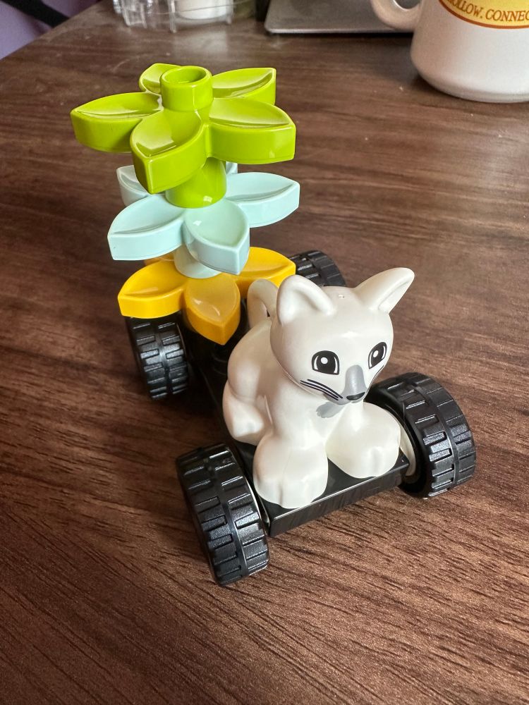 A small construction of Duplo blocks. A white cat and a stack of flowers on a set of wheels.