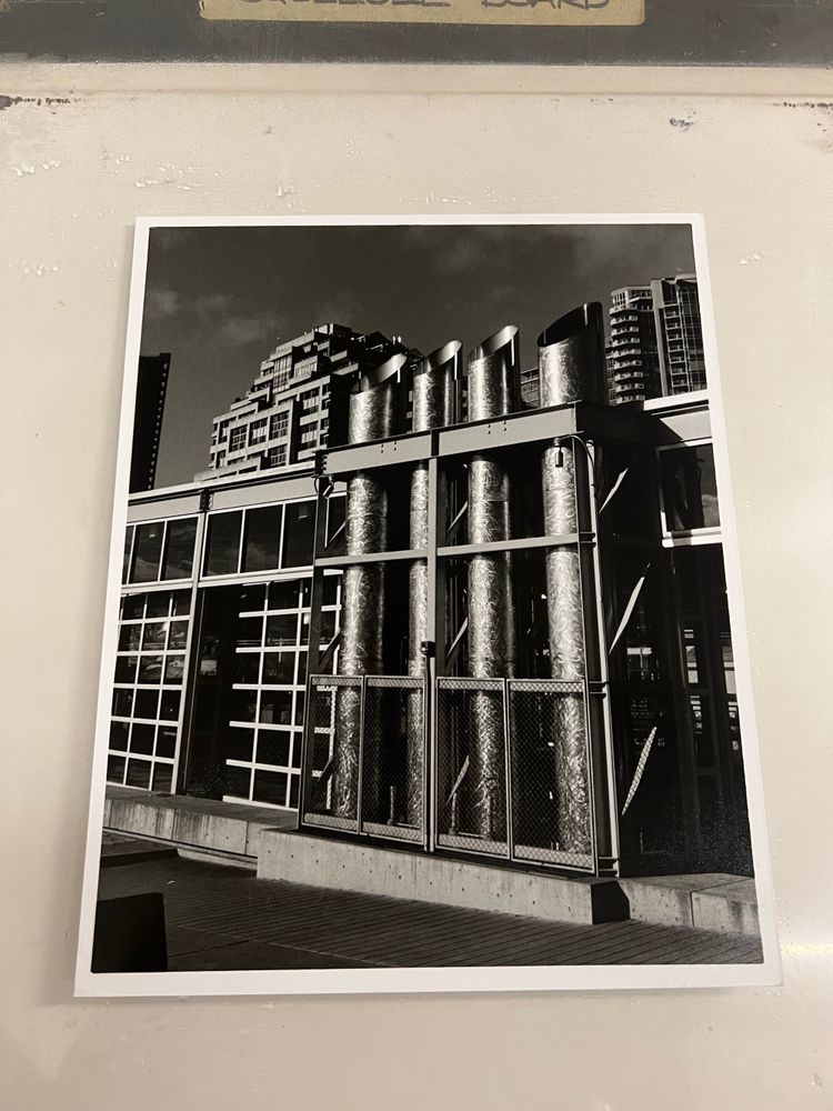 A black and white architectural composition, buildings with some smoke stacks in front. The smoke stacks glitter in the sunlight against the darker buildings and sky in the background