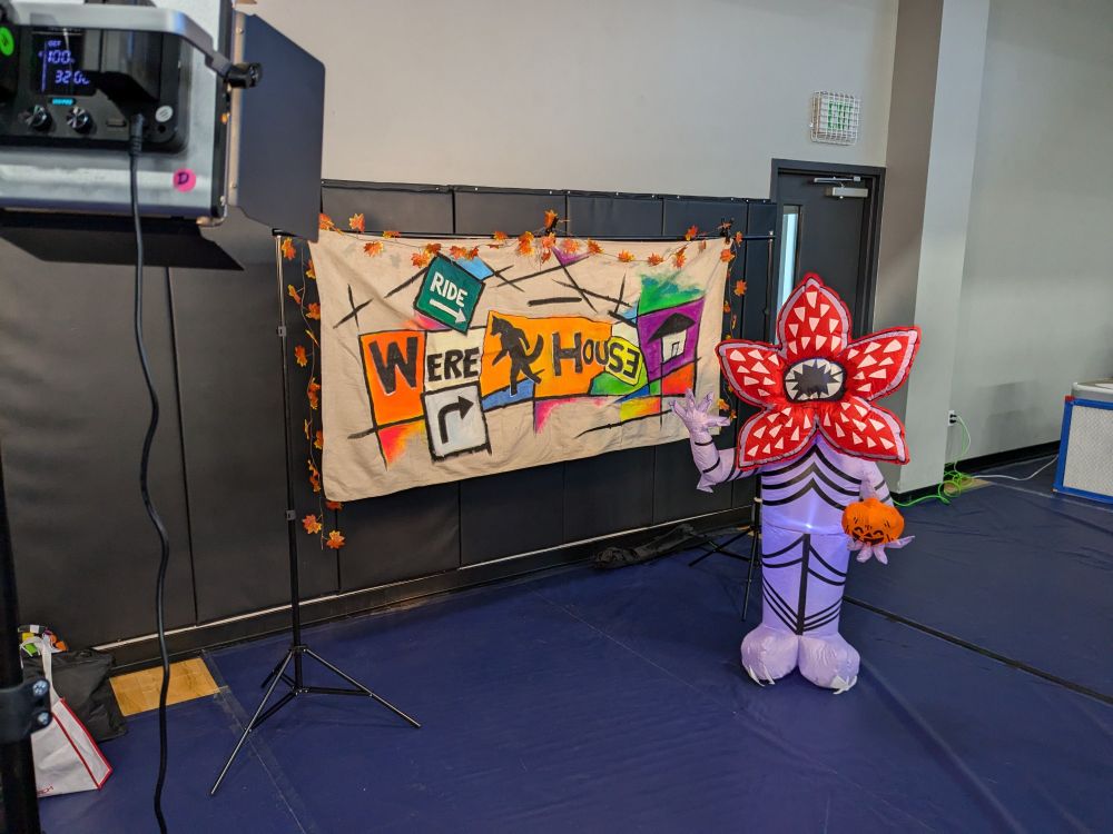 A photo area at last caw's were house art market with a colorful were house banner draped in fall leaves, with an inflatable monster called a demigorgon that kind of looks like a flower next to it 
