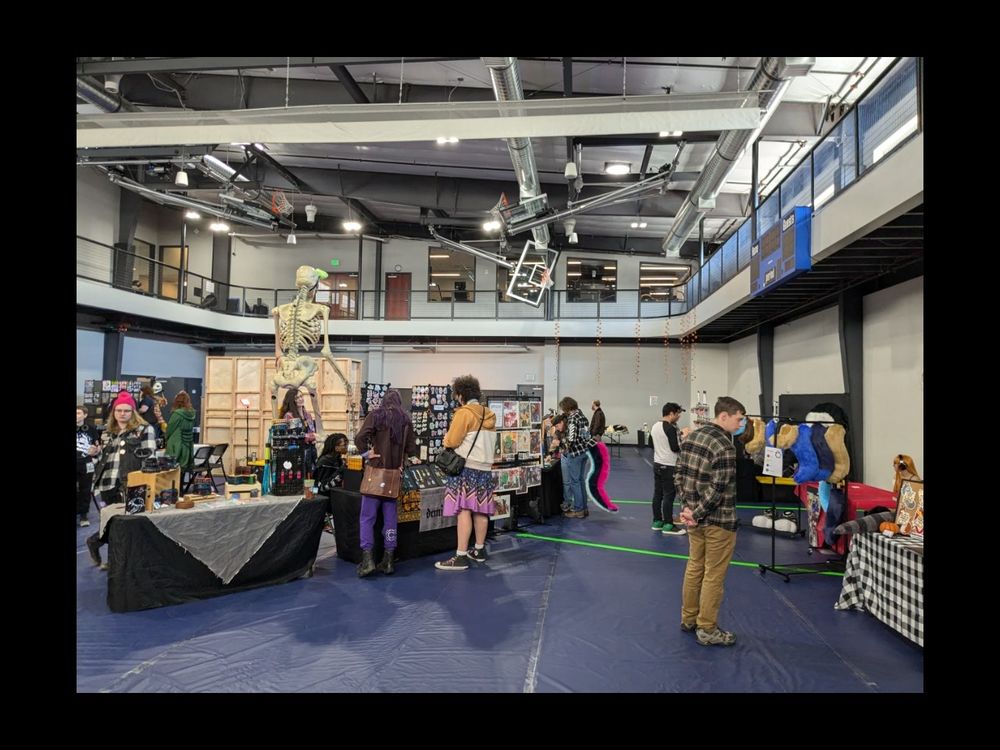 A gymnasium filled with art vendors and people perusing their wares. There is a giant skeleton in the middle of the room