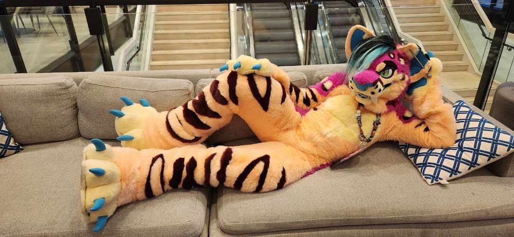 Saber tooth cat fursuit: beige, pink, blue - lounging on a cough. 