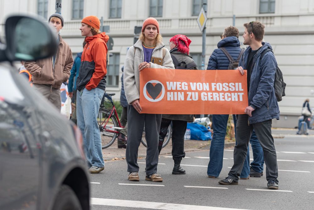 Mehrer Aktivist*innen, zwei halten ein Banner "Weg von Fossil hin zu gerecht". Im Vordergrund ein Auto.