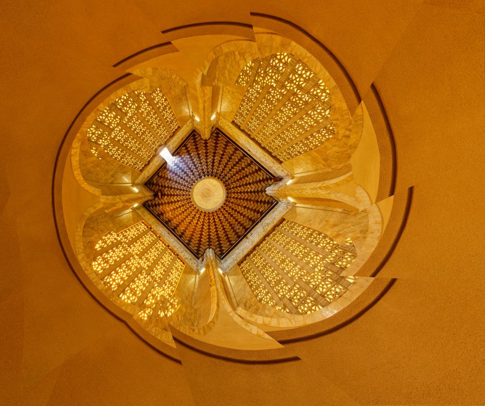 Most of the image is an brownish-orange colour. There is a broken circle on the outside of the square-shaped image. In the middle is a darker square, with pillars reaching up from the bottom to the top. The square has a light circle in the centre, and rays of darker tiles around the circle. 