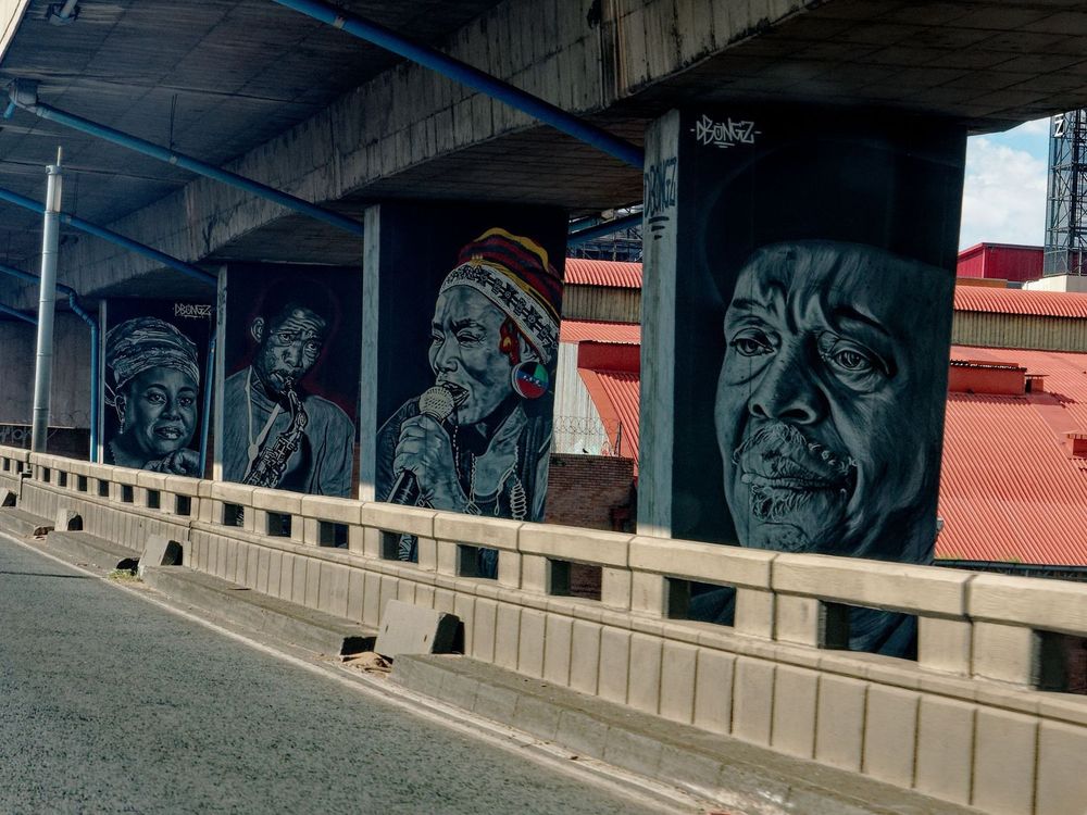 The scene shows the side of a road with concrete supports going to a second level of road above the first. On the supports are four faces, two men and two women. The woman and man in the middle of the four are making music.