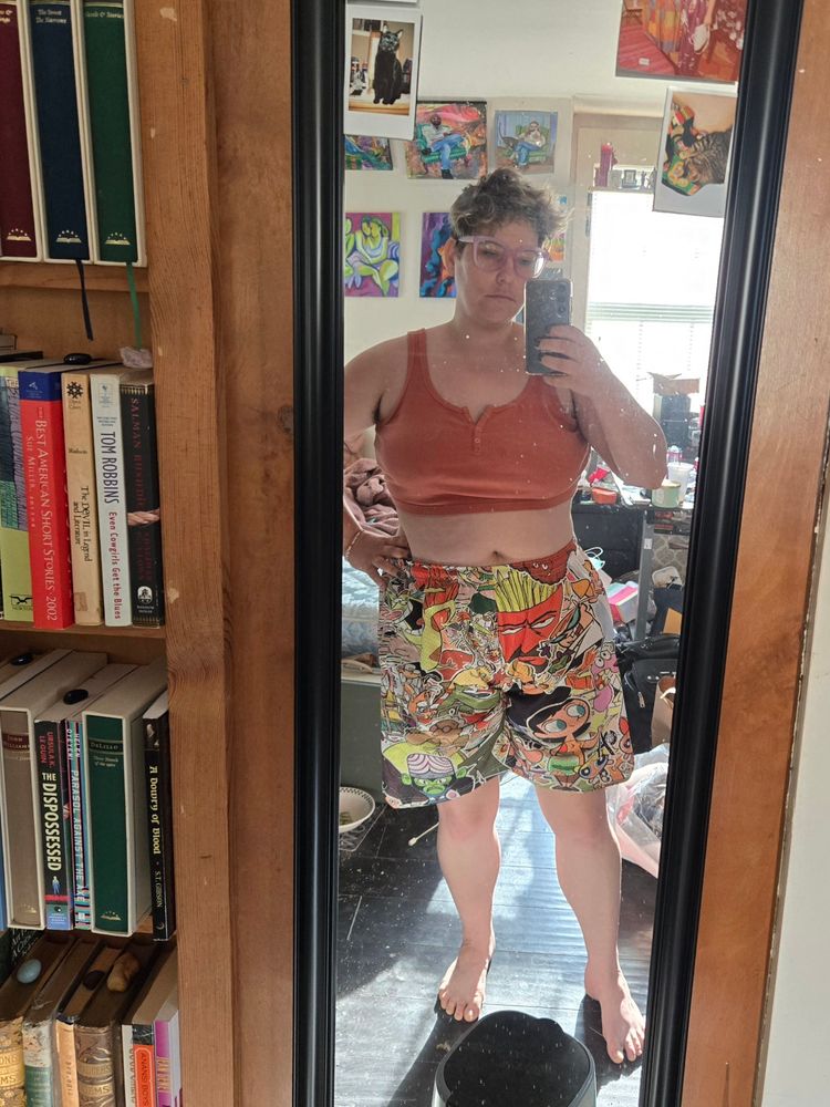 A mirror selfie of Kendall, a white person with short hair, wearing an orange crop tank and huge basketball shorts printed with Cartoon Network characters