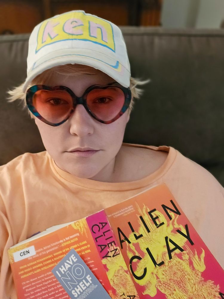 Kendall is sitting on a couch holding an open book and bookmark over her chest. He is wearing his custom "ken" baseball cap (that he painted in a Michael's parking lot), rose-tinted heart-shaped glasses, and a large peach t-shirt. The book is a paperback library book titled "Alien Clay" by Adrian Tchaikovsky. Her bookmark is blue and reads "I HAVE NO SHELF CONTROL" in bold font. 