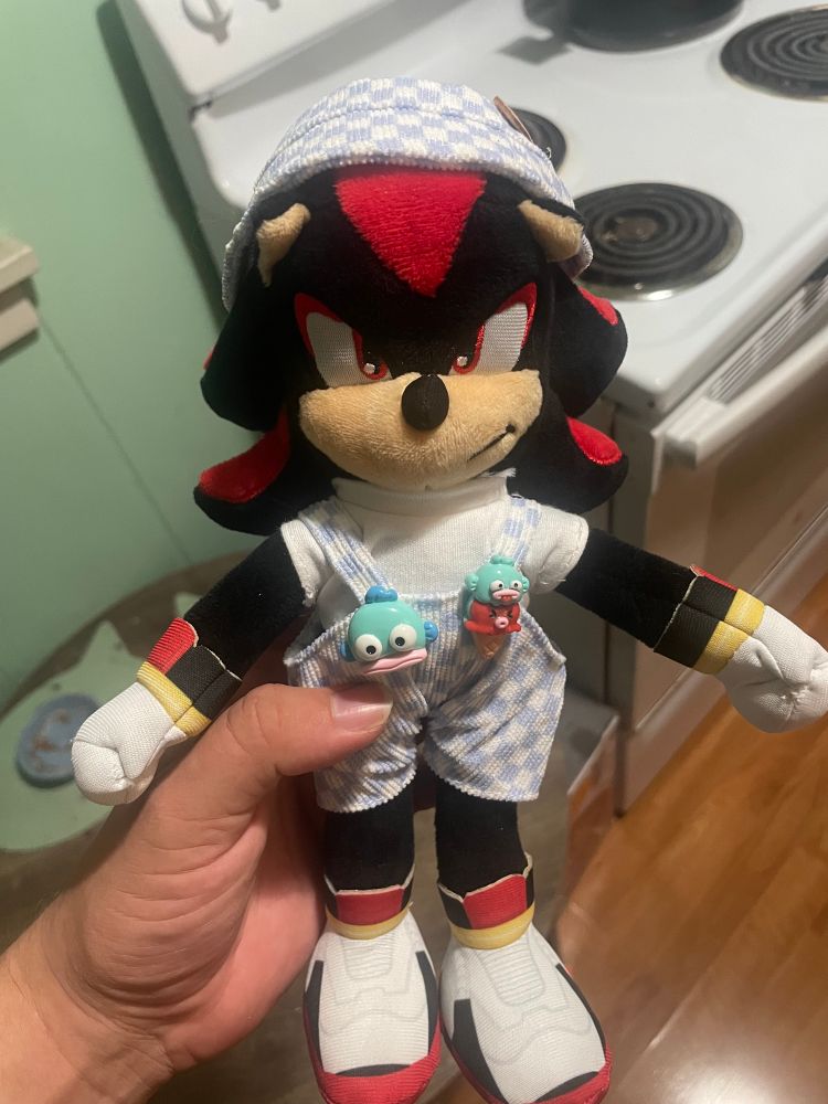 Shadow the hedgehog wearing a white shirt, corduroy overalls, and a bucket hat that have a light blue checkered pattern, on the overalls are two buttons