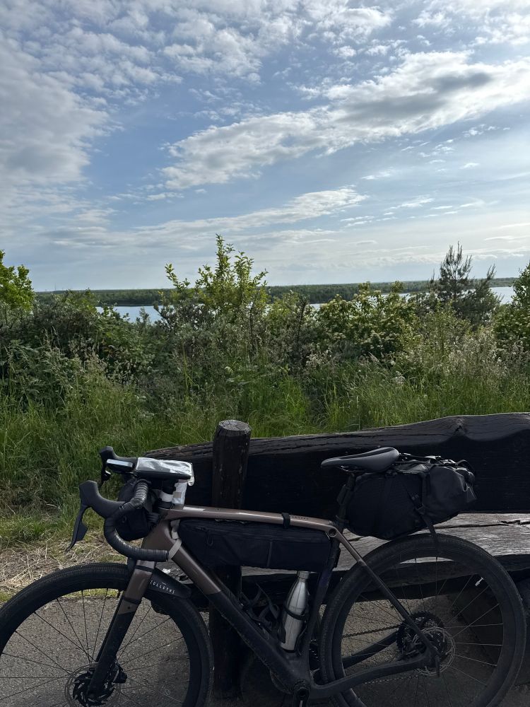 Im Vordergrund ein sehr dreckiges vollgepacktes Fahrrad, im Hintergrund Büsche und ein See