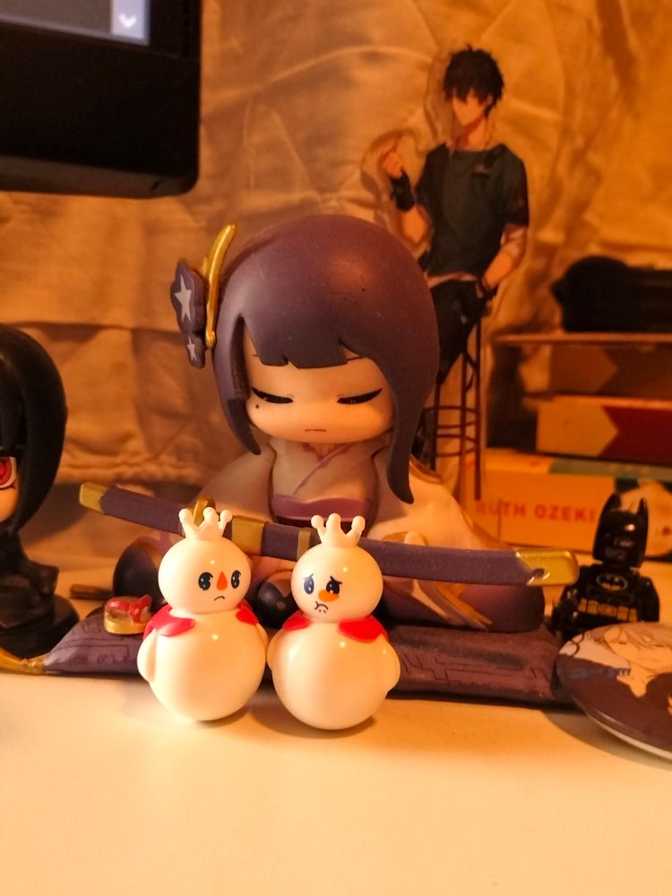 A desk picture. Two Mixue dolls, sitting infront of The Statue of Her Excellency The Almighty Narukami Ogosho God of Thunder. 