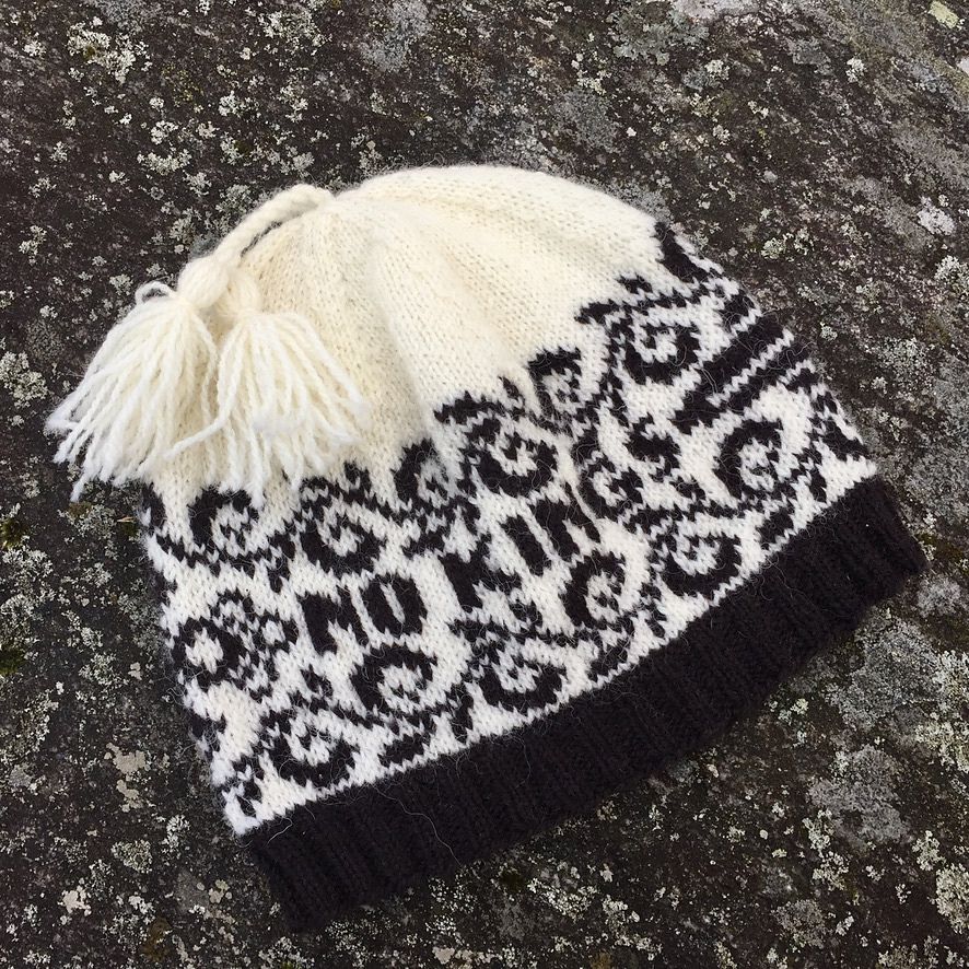 White hat with black design and lettering. NO KINGS is the central message surrounded by stylized black vines. The point of a sword comes in from the left, rest of sword wraps around hat. 