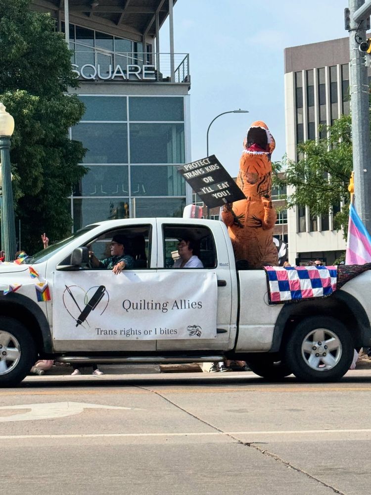A white pick up with a sign reading "Quilting Allies" and the slogan "Trans rights or I bites." In the bed of the pick up is a person in an inflatable T-Rex costume with a sign reading "Protect trans kids or I'll eat you!"