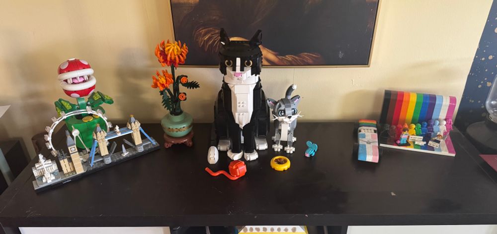 A bunch of Lego figures on top of a kallax shelf from ikea. London skyline, a piranha plant, some flowers, a big tuxedo cat next to a gray kitten and the everything is awesome set.