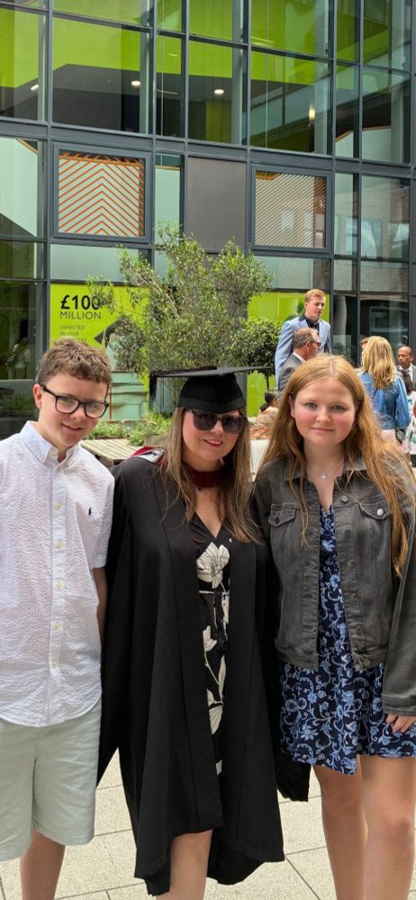 Image description: Three people standing outside a university building. Hazel is in the middle, wearing a black cloak and graduation cap. Her children stand either side of her. One boy and one girl.