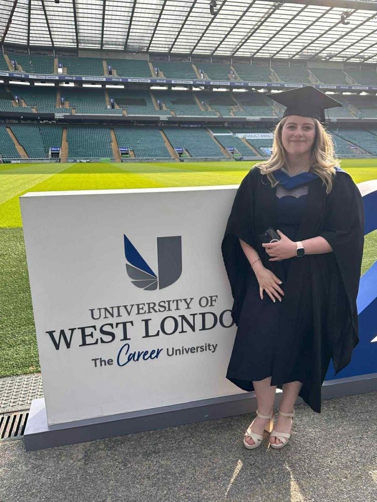 Image description: DSA member Katie stands inside Twickenham Stadium, wearing a graduation cloak and hat, next to a sign detailing "University of West London. The Career University ".