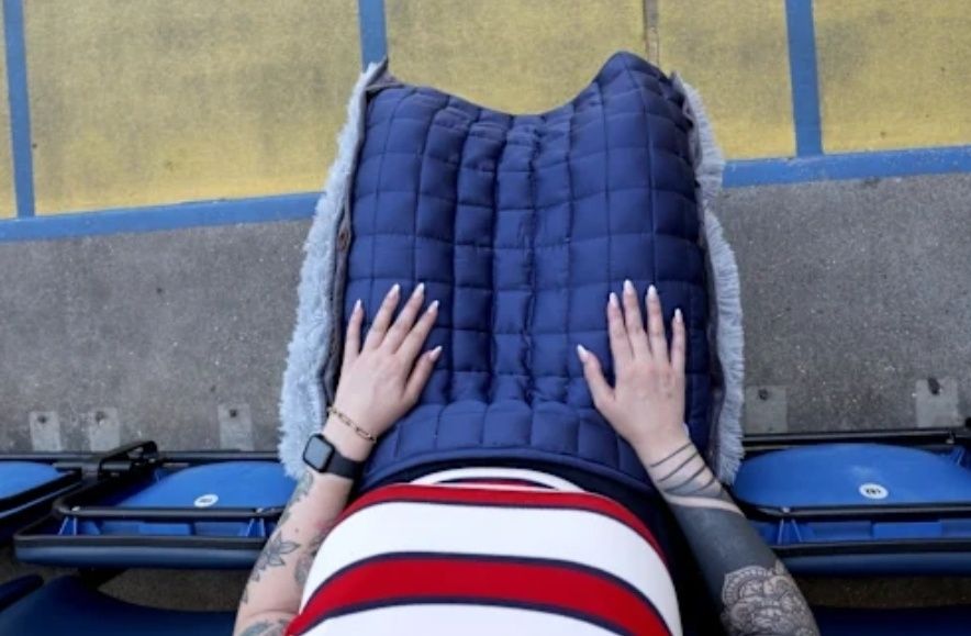 Image description: An overhead shot of a female, sat in a accessible seat at Stamford Bridge. A blue Zensory pad covers her thighs and knees.