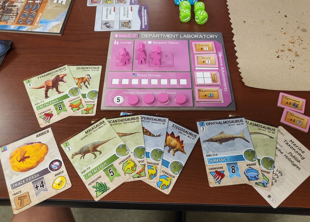 Cards and game pieces for the board game Holotype. There are discovery cards featuring ancient animals and a player board with researcher figures and tokens to track progress.