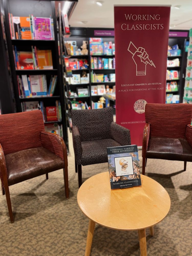 A photo taken before the book launch event showing three empty chairs and a table with the book A Periodic Table of Greek Mythology propped up on it. Behind the chairs is a roller banner for Working Classicists. 