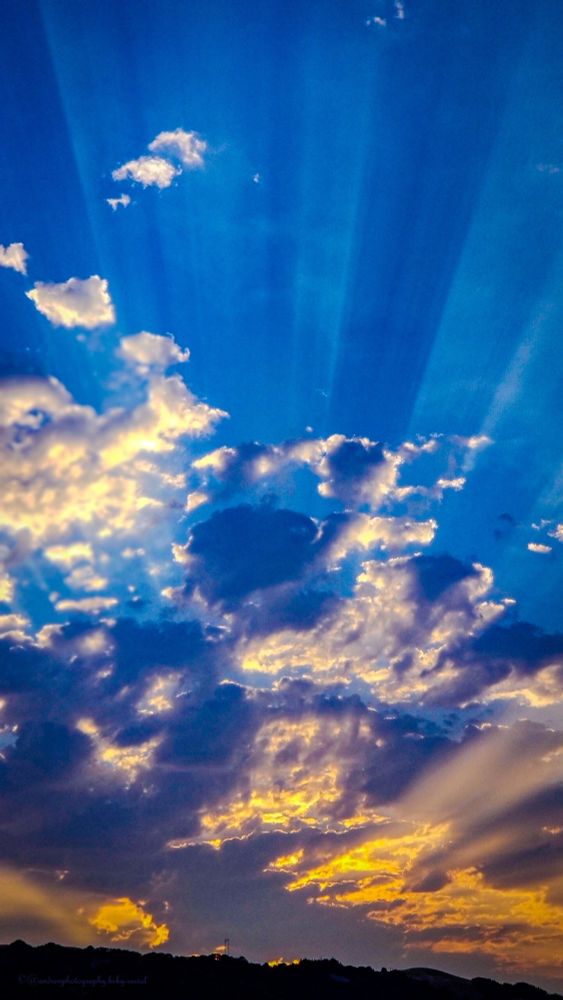 Dramatic sunset sky with golden crepuscular rays bursting through dark storm clouds over a silhouetted landscape. Brilliant beams of sunlight radiate across a deep blue sky, illuminating billowing clouds in shades of gold, orange, and gray above rolling hills.