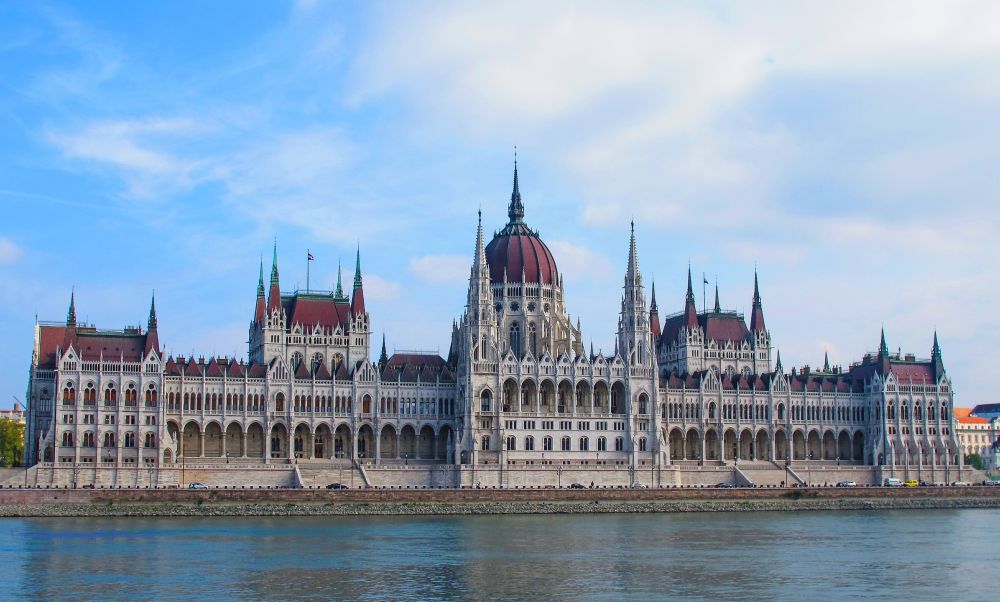 The majestic Hungarian Parliament Building in Budapest photographed from across the Danube River on a clear day. The Neo-Gothic architectural masterpiece features a prominent red dome flanked by symmetrical wings with intricate stone facades, pointed spires, and countless Gothic Revival details. The limestone building's pale exterior contrasts beautifully with its distinctive red-tiled roofs and copper dome, while the calm waters of the Danube reflect the structure under a partly cloudy blue sky