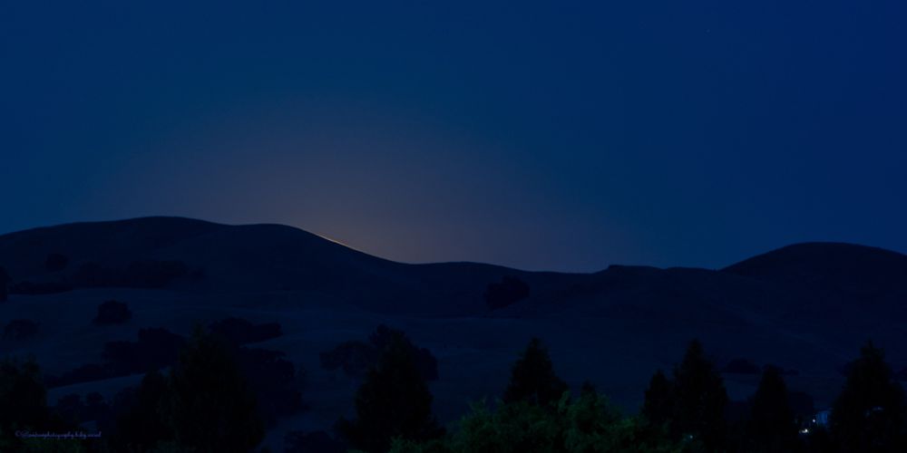 Twilight Anticipation:
Mystical twilight landscape with rolling mountain ridges silhouetted against a deep sapphire sky, dark evergreen trees dotting the foreground, and tiny golden lights sparkling in the distant valley like earthbound stars.