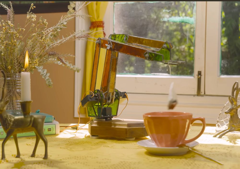 a servo arm with a hook sitting on a kitchen table, dunking a teabag into a cup of hot water. Only the arm is completely made of soldered stained glass.