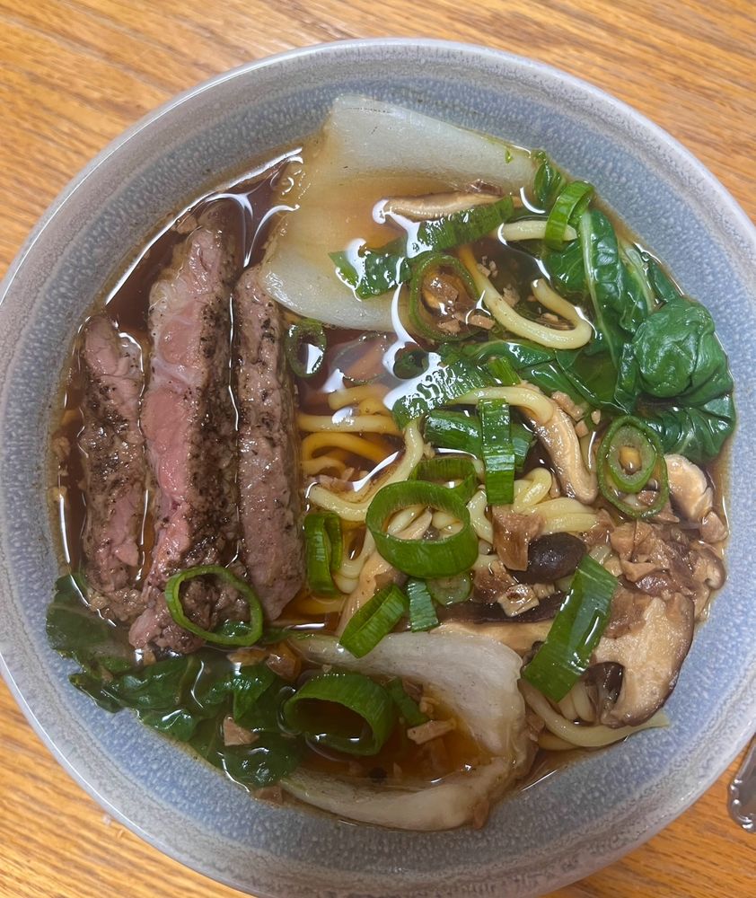 Ramen in a light blue bowl. Includes, ramen noodles, shiitake mushrooms, green onions, bok choy, steak, garlic, and broth