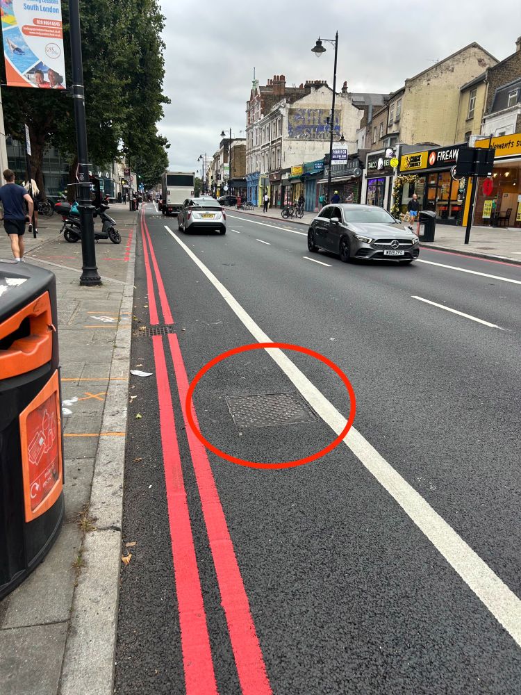 An image of CS7 on Clapham High Street in September 2025, with the same manhole cover highlighted for reference for the cycle lane width. The cycle lane has been reduced in width, with blue paint and wands removed.