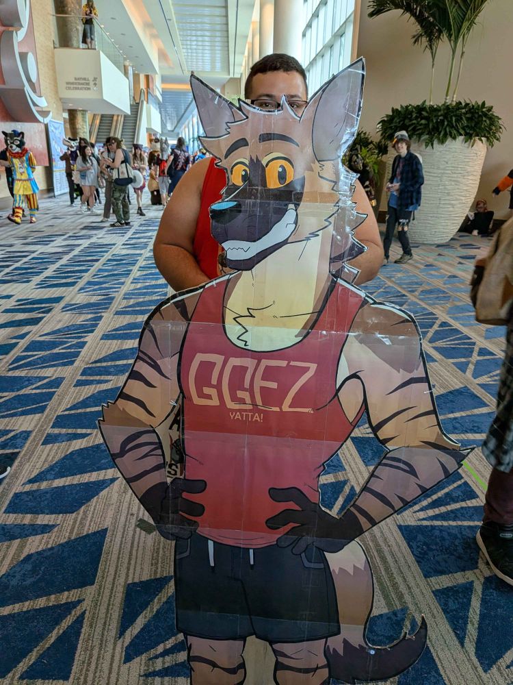 A cardboard cutout of Redline, a striped hyena man, wearing a red tank top that says GGEZ in yellow text. Redline is smiling wide with his hands on his hips. The cut out is in a hotel hallway during the furry convention Megaplex.