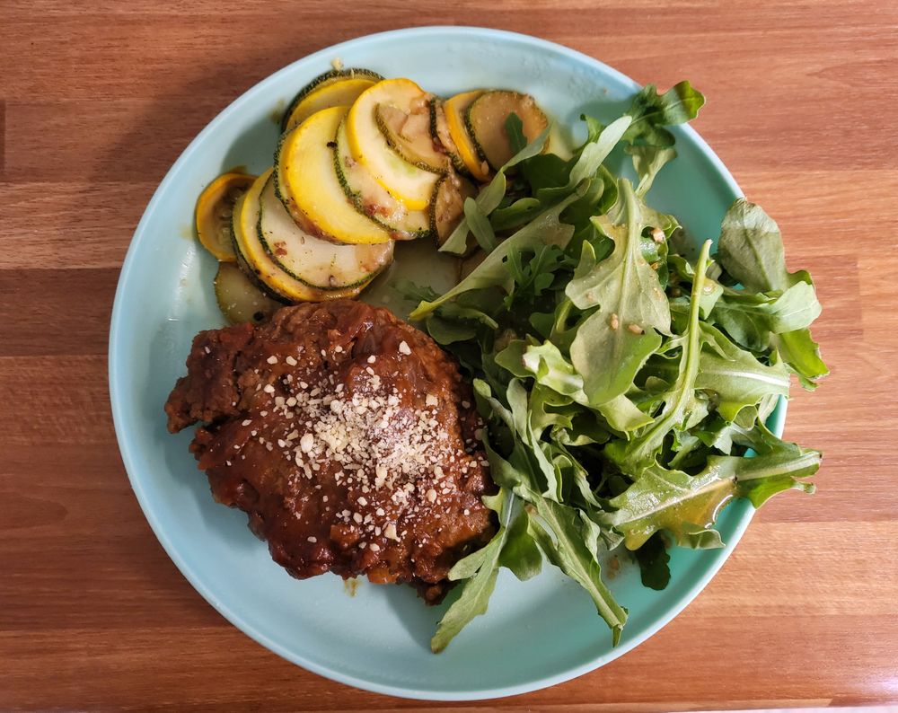 A small blue dish with arugula, thinly sliced zucchini, and a hefty beef patty. The beef patty is slathered in a dark sauce with caramalized onion bits, and topped with a dusting of parmesan cheese. The zucchini slices are stacked in alternating green and yellow slices, and has been broiled on top, melting the cheese and gently browning the edges of each slice. Some of the ponzu marinade has been drizzled over the argula like a thin dressing.