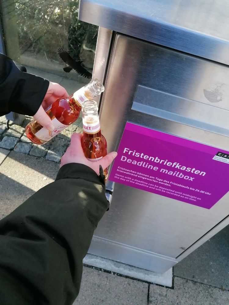 Links sind zwei Arme von jeweils einer anderen Person zu sehen, beide in schwarze Jacken eingekleidet. Sie stoßen sich zwei kleine Lillet White Peach-Flaschen zu. Im Hintergrund ist ein großer, silberner Briefkasten auf dem das magentapinke Schild steht: Fristenbriefkasten/Deadline mailbox. 
