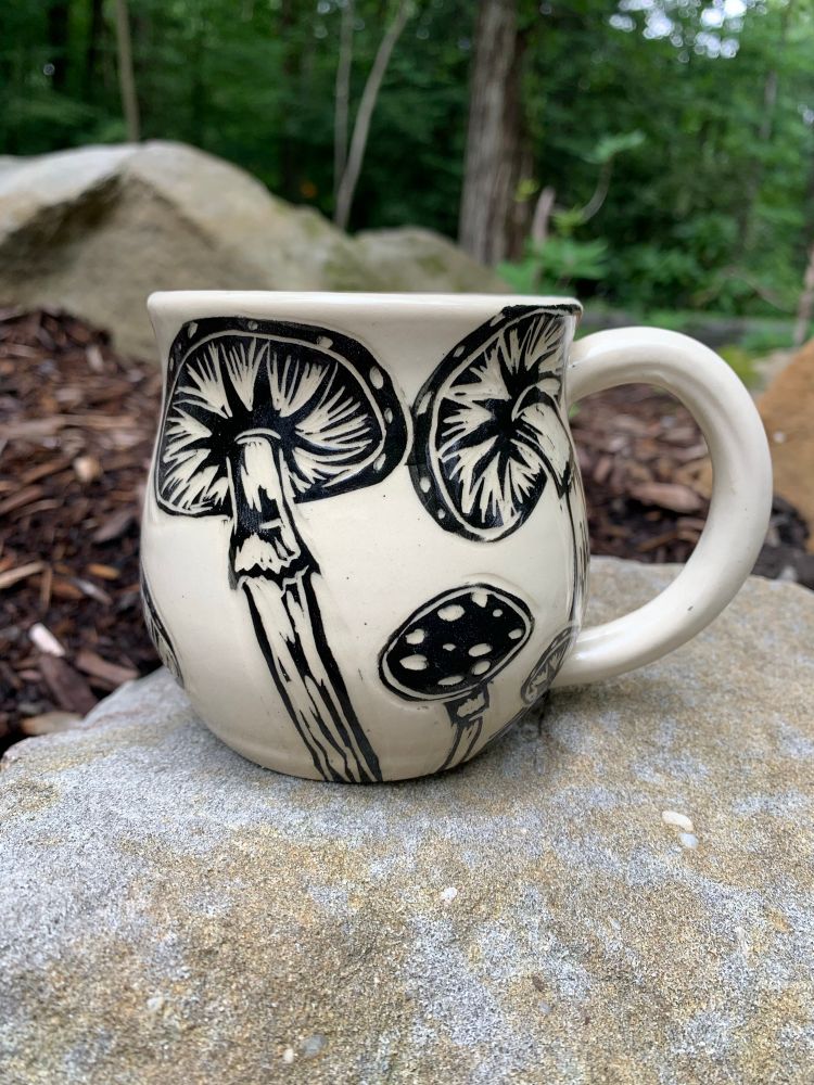 A mug, sitting on a rock in nature. The handle is to the right, showing the back of the mug. The mug has a blue interior. The surface is decorated using sgraffito, and from this angle you can see the image of large mushrooms. 