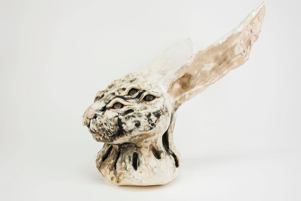 A ceramic sculpture of a the head of a creature resembling a white hare, facing left. It has 3 eyes visible: 6 total on the sculpture. The clay is off-white, with patches of brighter white in some places where slip was placed. The sculptures details were rubbed with black, allowing the fur to be seen. The nose, ear interiors, and under-eyes have a very slight pink glaze. The eyes themselves are deep-set, and glazed with an antique gold glaze: it is darker where thin (around the eye) and gold where thick, which is where the iris is. The sculpture comes to an end at the lower neck area of the hare. 