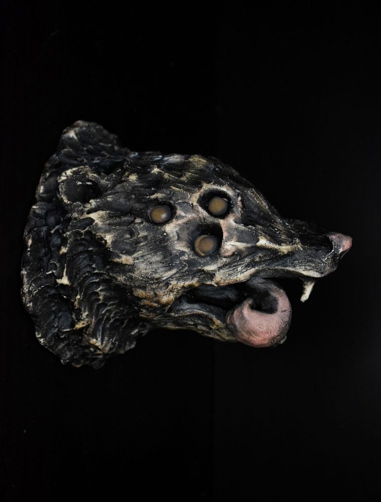 A ceramic sculpture in the form of the head of a creature inspired by a possum, facing right. Its tongue is pink and curled downward back into itself. It has a pink nose, and one white fang is showing, as well as the side of a pink nose. It features three eyes: glazed with antique bronze glaze, which fades from a black to a muted gold in the center. 