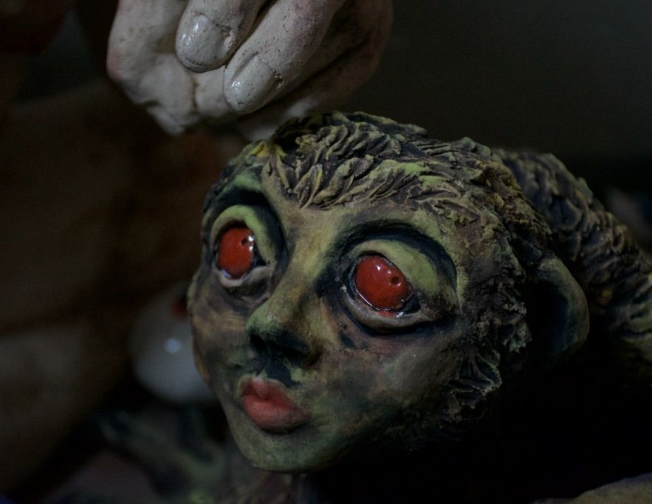 The face of a sculpture, in a dark setting surrounded by other sculptures. The face has large red eyes (resembling those of a bush baby), a humanoid nose and lips, and round ears on the side of its head. It is furry, the fur a dirty greenish yellow. This is a side view of the face. 