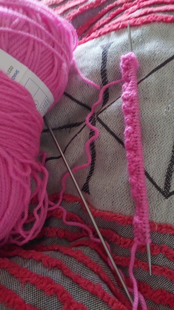 Pink DK acrylic ball of wool on the left, two DP needles on the right, one of which has three rows of wobbly ribbing on it.