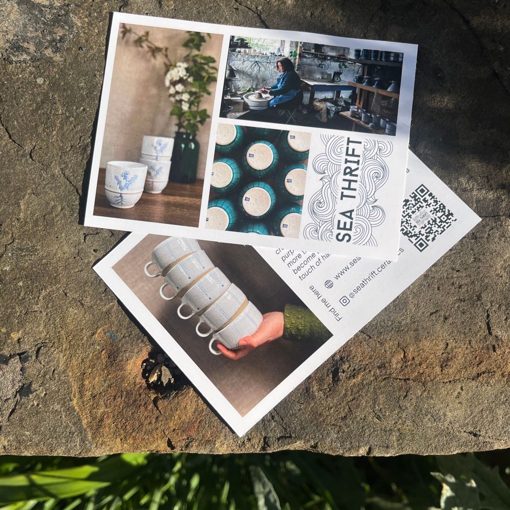 Flyers on a stone wall with pictures of a stack of handmade cups balanced in a hand, a woman with curly hair and a blue apron working at a pottery wheel in a ceramics studio, a white ceramic wine cup with a dandelion print in blue, a logo with the title ‘seathrift’