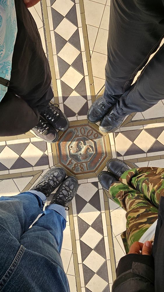 Four pairs of feet as seen from above, standing at even intervals around a metal medallion embedded in the floor. The medallion says 1884 on one side and 2009 on the other with a twin fish emblem in the middle. The floor around it is white and black tiling in diamond shapes.