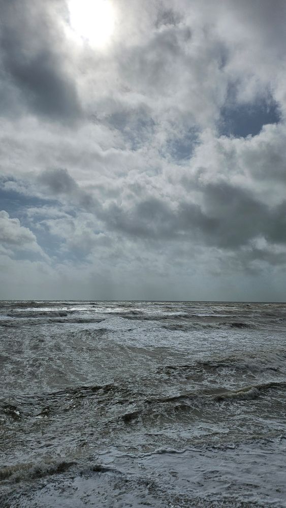 A choppy, grey-brown sea with a cloudy sky that threatens a storm to come. The sun can be seen illuminating the back of the clouds.