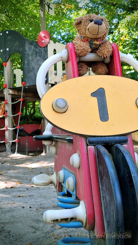 FOTO eigen

Das Bild zeigt Bärnhardt, den Lümmelteddy, der auf einem Spielplatzgerät sitzt.

Der Teddybär ist flauschig, hat ein braunes Fell und trägt einen bunten, gestreiften Schal um den Hals. Er sitzt auf einem Federwippgerät, das wie ein Motorrad gestaltet ist.

An dem Motorrad befindet sich eine gelbe, ovale Platte mit der schwarzen Ziffer "1" darauf.

Im Hintergrund sind weitere Elemente eines Spielplatzes zu erkennen, darunter ein Kletternetz und grüne Bäume, die viel Laub tragen. Der Boden des Spielplatzes besteht aus Erde oder Sand. 

Die Szene ist bei Tageslicht aufgenommen.
