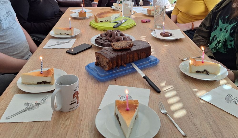 Ein gedeckter Tisch für 8 Personen. In der Mitte stehen zwei Kuchen und ein Teller mit Lebkuchen und Dominosteinen. Um den Tisch sieht man Teile von Personen, ohne Köpfe, da diese aus dem Bildausschnitt herausragen.
Es stehen 8 Teller auf dem Tisch, 7 mit je einem Stück Käsekuchen, 1 mit einem Lebkuchen.
In jedem Stück steckt eine brennende Geburtstagskerze, bereit, ausgepustet zu werden.