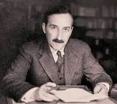 Ein Foto von Stefan Zweig am Schreibtisch. Er hält einen Brief in der Hand und schaut den Betrachter an.