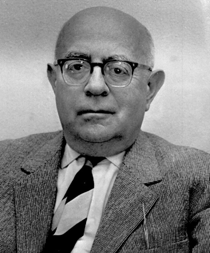 Ein Foto von ihm. Er trägt einen Anzug mit Hemd und Krawatte, darüber ein breites, älteres Gesicht mit einer Brille.